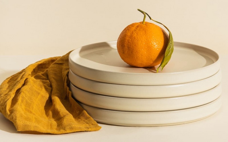 lifestyle image of a stack of plates with an orange on top and a orange towel beside it